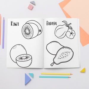 May include: A black and white coloring book page with illustrations of kiwi and papaya fruits. The page is open on a white surface with colored pencils and small triangular blocks.