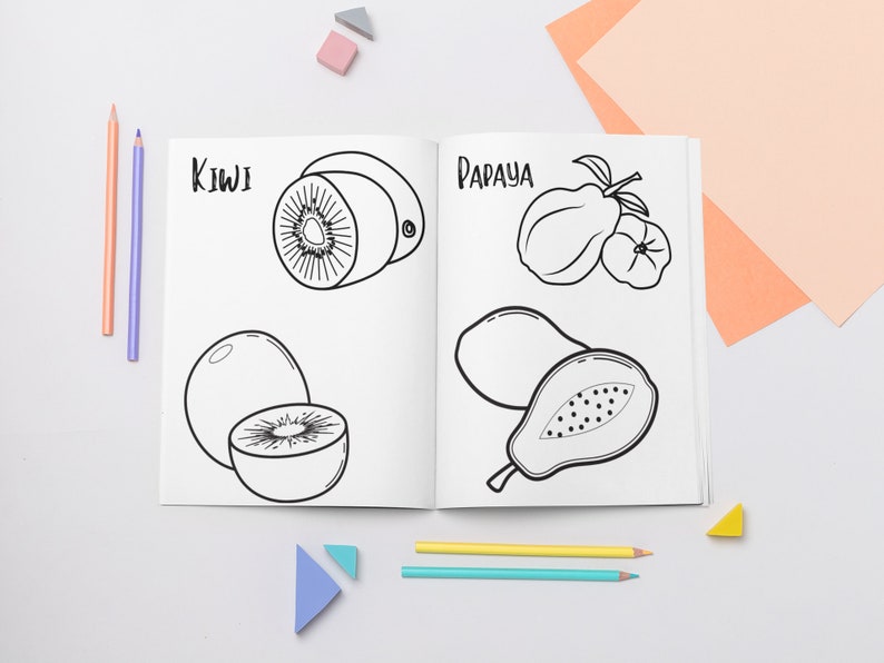May include: A black and white coloring book page with illustrations of kiwi and papaya fruits. The page is open on a white surface with colored pencils and small triangular blocks.