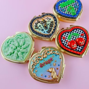May include: Five heart-shaped gold-tone compact mirrors with decorative lids. One features green mice, one says 'dead inside' with a skeleton, one says 'CREATURE COMFORT' with a green monster, one has red cherries, and one says 'drugs' with a gold bird.