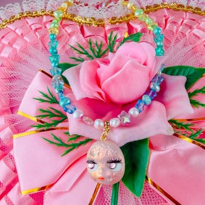 May include: A beaded necklace featuring a mix of small white pearls, translucent blue star-shaped beads, and yellow beads. A round, pale pink pendant with a stylised face, including dark eyelashes and pink lips, hangs from a gold-toned metal bail.