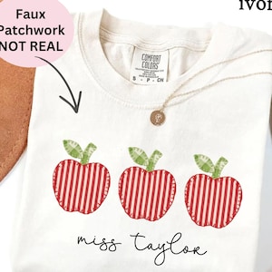 Pode incluir: Camiseta cor marfim com três desenhos de maçãs com padrões de listras vermelhas e brancas e folhas verdes. O texto "miss taylor" está escrito em itálico abaixo. Um colar de ouro está sobre a camiseta.