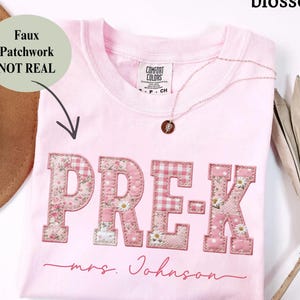 Op de afbeelding: Een lichtroze t-shirt met het woord "PRE-K" in een patchwork-ontwerp. De letters zijn een mix van bloemen- en ruitpatronen. De tekst "mrs. Johnson" staat eronder in cursief. Een gouden ketting met een hanger ligt op het shirt.