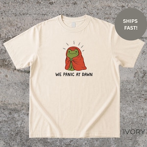 以下が含まれることがあります： アイボリー色のTシャツ。赤い毛布に包まれた漫画のカエルが描かれています。カエルは心配そうな表情をしています。「WE PANIC AT DAWN」の文字が下にあります。Tシャツは半袖で、クルーネックです。