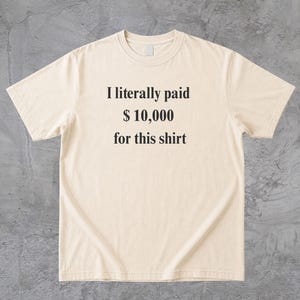 Pode incluir: Camiseta creme com o texto "I literally paid $10,000 for this shirt" em preto. A camiseta tem mangas curtas e gola redonda. O tecido parece ser uma mistura macia de algodão.