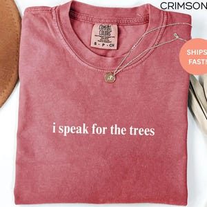 May include: Crimson-colored t-shirt with the phrase "i speak for the trees" in white lettering. The shirt has a crew neck and a small tag on the upper chest. A silver necklace with a pendant is draped across the shirt.
