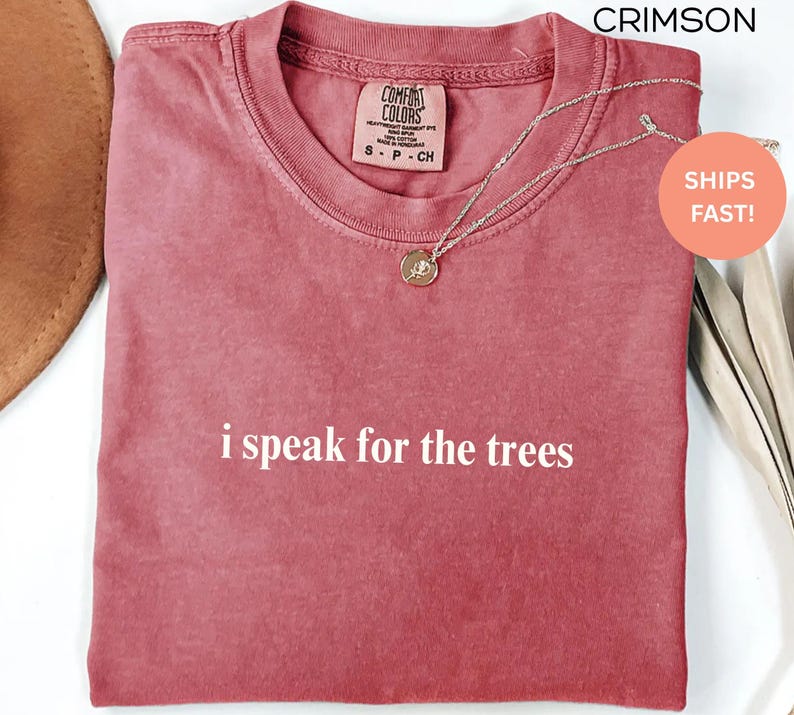May include: Crimson-colored t-shirt with the phrase "i speak for the trees" in white lettering. The shirt has a crew neck and a small tag on the upper chest. A silver necklace with a pendant is draped across the shirt.