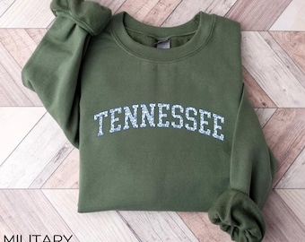 Op de afbeelding: Militairgroen sweatshirt met het woord "TENNESSEE" in een wit en blauw bloemmotief. Het sweatshirt is op een houten oppervlak gevouwen. De tekst "MILITARY GREEN" staat linksonder in de afbeelding.