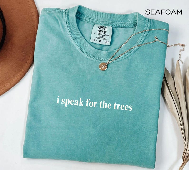 May include: Seafoam green t-shirt with the phrase "i speak for the trees" in white. A gold necklace with a pendant rests on the shirt. The shirt is a Comfort Colors brand.