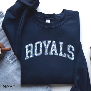 May include: Navy blue sweatshirt with the word "ROYALS" in white letters with a blue floral pattern. The word "NAVY" is printed at the bottom left of the image. The sweatshirt is made of a soft material.