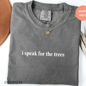 May include: A gray t-shirt with the text "i speak for the trees" in white. The shirt has a crew neck and short sleeves. A silver necklace with a circular pendant is on the shirt.