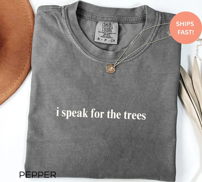May include: A gray t-shirt with the text "i speak for the trees" in white. The shirt has a crew neck and short sleeves. A silver necklace with a circular pendant is on the shirt.