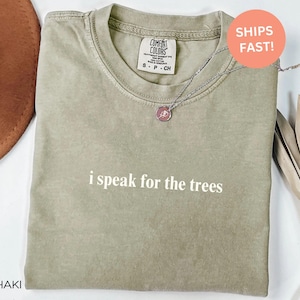 May include: Khaki-colored t-shirt with the phrase "i speak for the trees" in white. A silver necklace with a circular pendant is on the shirt. The shirt has a Comfort Colors tag. A brown hat is partially visible.