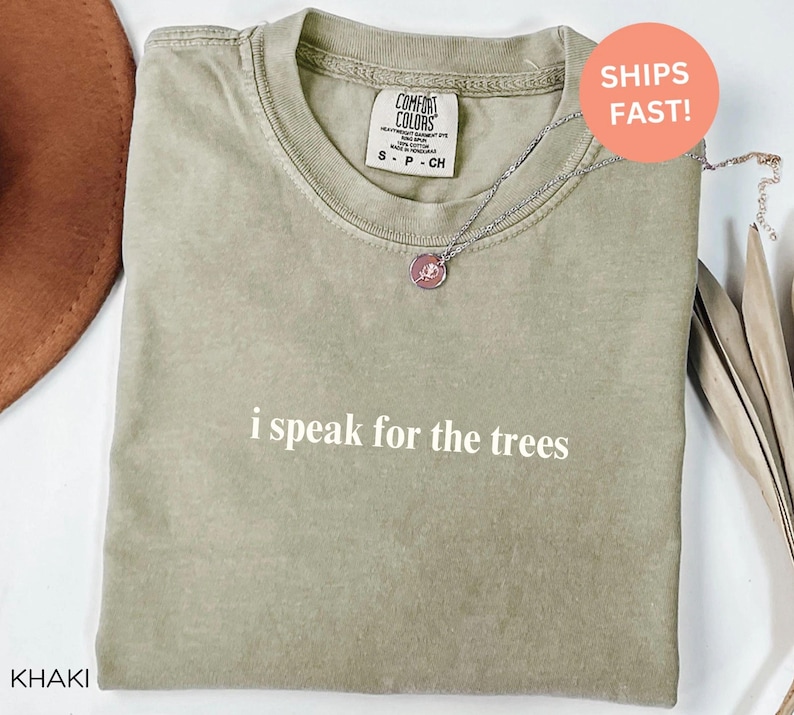 May include: Khaki-colored t-shirt with the phrase "i speak for the trees" in white. A silver necklace with a circular pendant is on the shirt. The shirt has a Comfort Colors tag. A brown hat is partially visible.