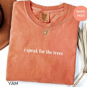 May include: A coral-colored t-shirt with the phrase "i speak for the trees" printed in white. The shirt has a crew neck and short sleeves. A gold necklace with a pendant is draped over the shirt. The tag reads "Comfort Colors".