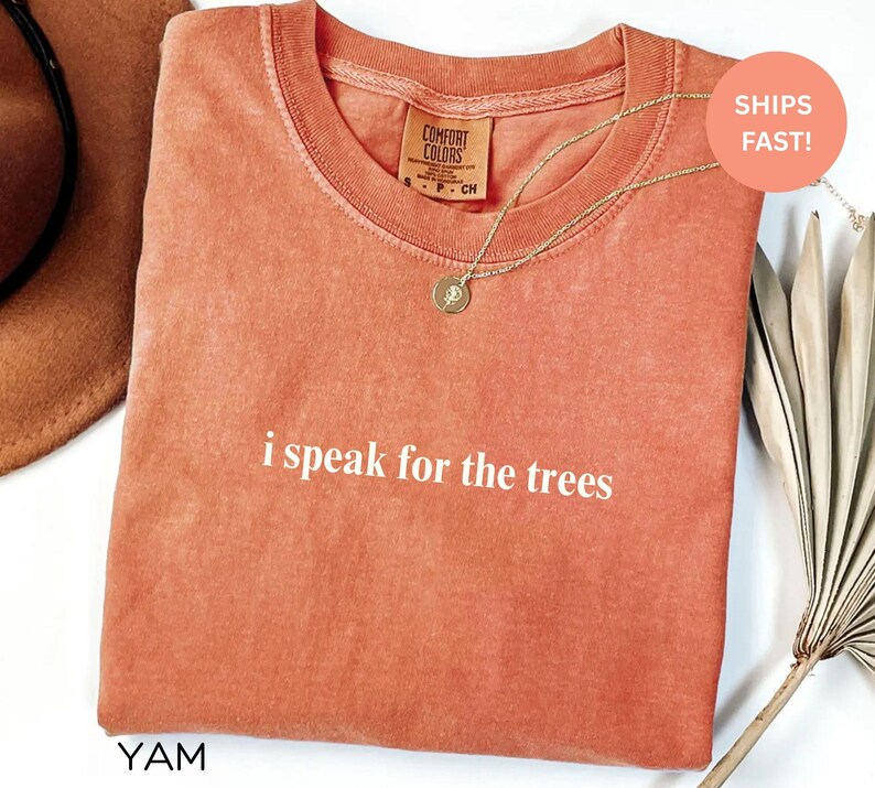 May include: A coral-colored t-shirt with the phrase "i speak for the trees" printed in white. The shirt has a crew neck and short sleeves. A gold necklace with a pendant is draped over the shirt. The tag reads "Comfort Colors".