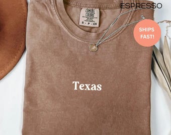 Puede incluir: Camiseta color espresso con la palabra "Texas" impresa en blanco. La camiseta está doblada y tiene una etiqueta Comfort Colors. Un collar plateado con un pequeño colgante está sobre la camiseta.
