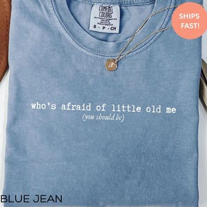May include: A blue jean colored t-shirt with the text "who's afraid of little old me (you should be)". A gold necklace with a pendant is draped across the shirt. The shirt has a Comfort Colors tag.