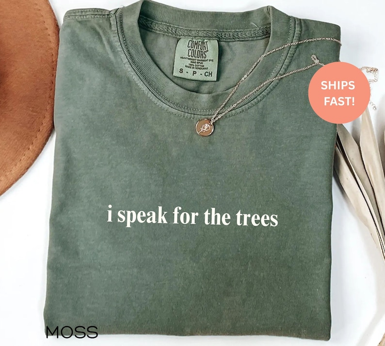 May include: Moss green t-shirt with the text "i speak for the trees" in white. The shirt has a crew neck and short sleeves. A silver necklace with a circular pendant is on the shirt.