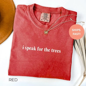 May include: Red t-shirt with the phrase "i speak for the trees" in white lettering. A gold necklace with a pendant rests on the shirt. The shirt has a crew neck and a tag that says "Comfort Colors".