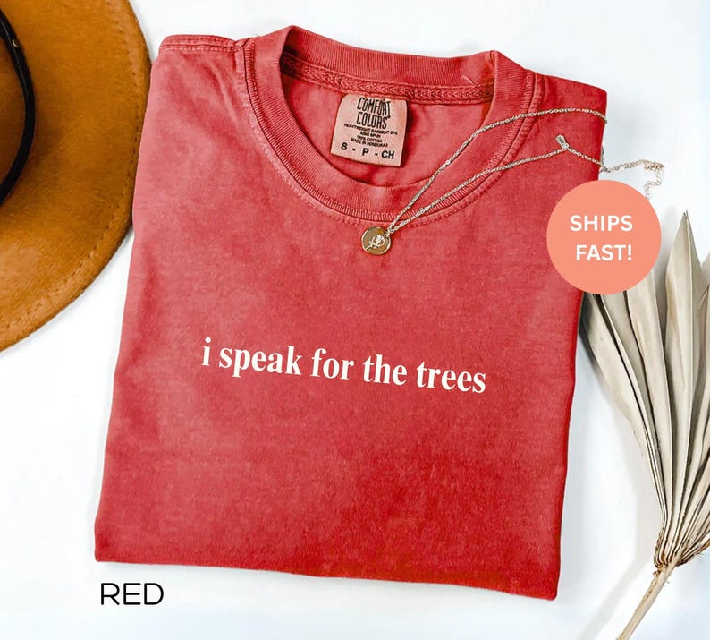 May include: Red t-shirt with the phrase "i speak for the trees" in white lettering. A gold necklace with a pendant rests on the shirt. The shirt has a crew neck and a tag that says "Comfort Colors".