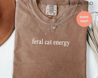 Puede incluir: Camiseta color espresso con la frase "feral cat energy" en blanco. La camiseta tiene cuello redondo y mangas cortas. Un collar plateado con un pequeño colgante está sobre la camiseta.