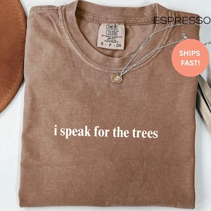 May include: Espresso-colored t-shirt with the text "i speak for the trees" in white. The shirt has a crew neck and short sleeves. A silver necklace with a pendant is draped across the shirt.