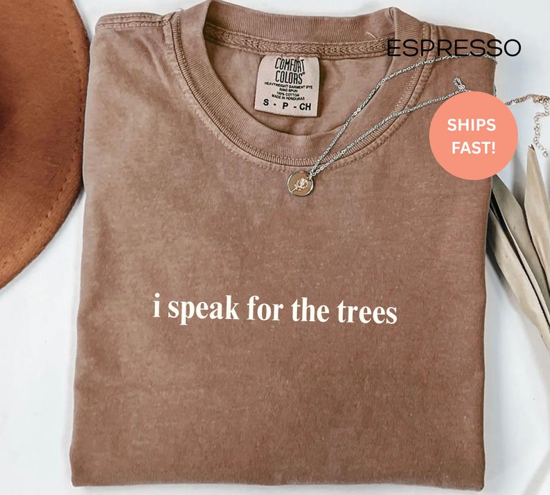 May include: Espresso-colored t-shirt with the text "i speak for the trees" in white. The shirt has a crew neck and short sleeves. A silver necklace with a pendant is draped across the shirt.