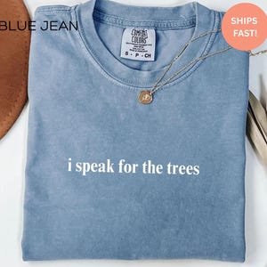 May include: A blue t-shirt with the text "i speak for the trees" in white. The shirt has a crew neck and short sleeves. A gold necklace with a pendant is on the shirt.