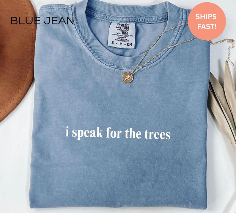 May include: A blue t-shirt with the text "i speak for the trees" in white. The shirt has a crew neck and short sleeves. A gold necklace with a pendant is on the shirt.