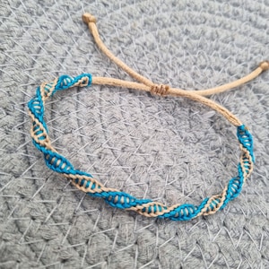 Op de afbeelding: Een handgemaakte armband met een geweven ontwerp. De armband heeft een beige koord met een blauw en beige geweven patroon. De verstelbare sluiting is beige. De armband ligt op een grijs geweven oppervlak.