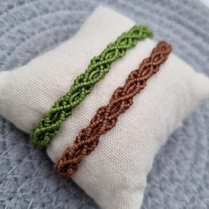 以下が含まれることがあります： 2つのマクラメブレスレット、緑と茶色が、小さな白い枕の上に置かれています。ブレスレットは織り込まれたデザインです。背景は明るい灰色のテクスチャ加工された表面です。画像は明るく、ブレスレットの細部を際立たせています。