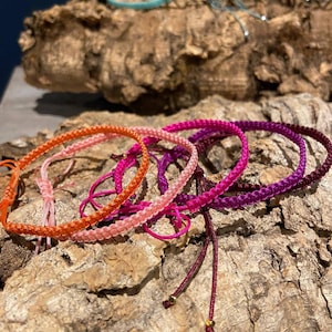 Op de afbeelding: Een set kleurrijke gevlochten armbanden met gouden kralen. De armbanden zijn gemaakt van verschillende kleuren, waaronder roze, oranje, rood en paars. De armbanden zijn op een stuk hout gestapeld.