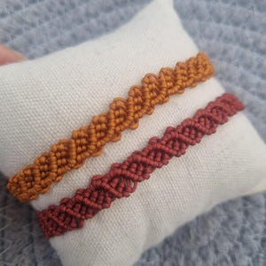 以下が含まれることがあります： オレンジと赤褐色の2色のマクラメブレスレットが白い枕の上に置かれています。ブレスレットは織り模様です。枕は灰色の表面にあります。画像には「B❤hemianNL」というテキストが含まれています。