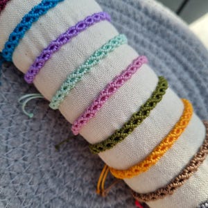 Op de afbeelding: Een verzameling handgemaakte macramé-armbanden in verschillende kleuren, waaronder paars, blauw, groen, roze en bruin. De armbanden worden getoond op een witte stoffen rol, die hun ingewikkelde geweven ontwerp laat zien. De armbanden zijn verstelbaar.