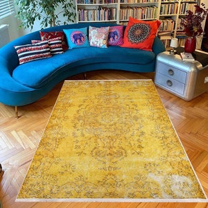 May include: A vintage yellow rug with a floral pattern and faded areas. The rug is on a wooden floor in a living room with a blue sofa and cushions.