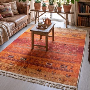 Puede incluir: Un salón acogedor con un sofá de cuero marrón, una vibrante alfombra naranja y una estantería de madera llena de libros. Plantas en macetas se encuentran en un banco de madera cerca de una gran ventana. Objetos decorativos y obras de arte enmarcadas adornan las paredes.