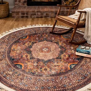 May include: A circular rug featuring an intricate pattern in brown, orange, and blue hues. It has a cream-coloured border with tassels. The rug is positioned in front of a fireplace, with a rocking chair nearby.