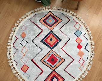 May include: A circular rug in light grey, featuring a geometric diamond design in red, orange, blue, and navy. It has a cream-coloured border with tassels. The rug is on a wooden floor.