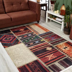 Op de afbeelding: Een veelkleurig patchwork tapijt met verschillende geometrische patronen in tinten rood, oranje, bruin, zwart, wit en groen. Het tapijt heeft een witte rand en ligt op een lichte houten vloer.