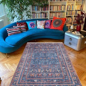 May include: A blue rug with a geometric pattern featuring stylized animals and plants. The rug has a white fringe border and is laid out on a hardwood floor in front of a blue sofa with colorful throw pillows.