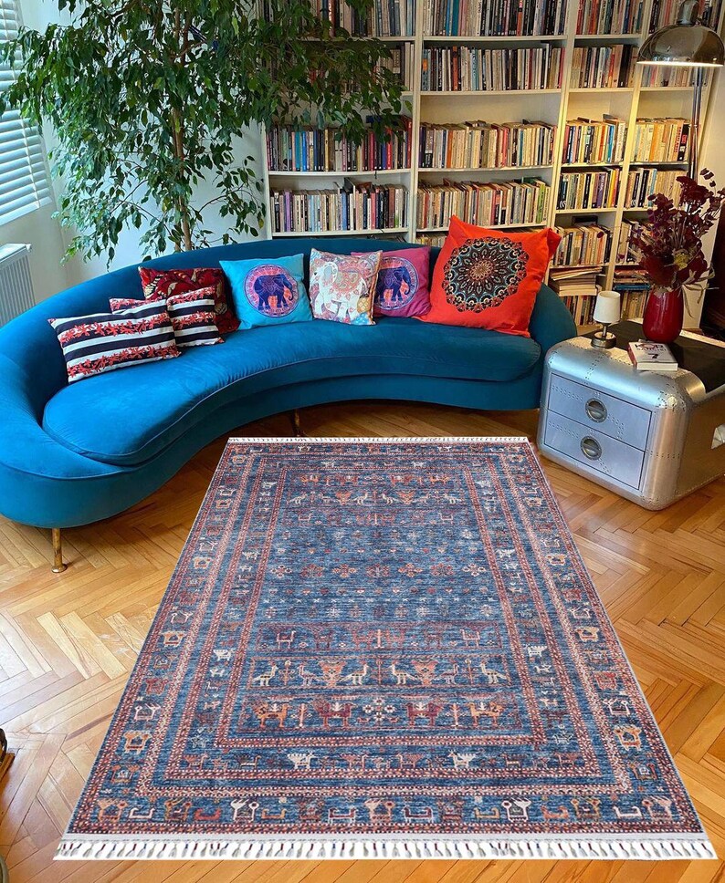 May include: A blue rug with a geometric pattern featuring stylized animals and plants. The rug has a white fringe border and is laid out on a hardwood floor in front of a blue sofa with colorful throw pillows.