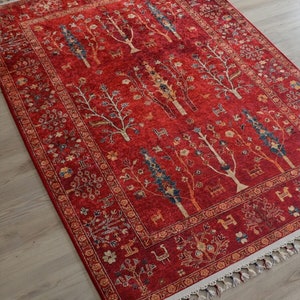 May include: A red rectangular rug with a detailed tree and animal design. The rug has a border with floral patterns and a fringe along the edges. The trees are white and blue, and the animals are brown.