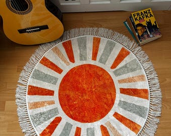 May include: Round rug with a sunburst design. The rug features a vibrant orange center with radiating stripes in orange, gray, and white. It has a fringed border and is placed on a wooden floor. A guitar and books are in the background.