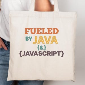 May include: A cream-coloured tote bag featuring the text "Fueled by Java {&} JavaScript" in orange, green, yellow, and brown. The bag has two handles and is held by a person wearing a white t-shirt and blue jeans.