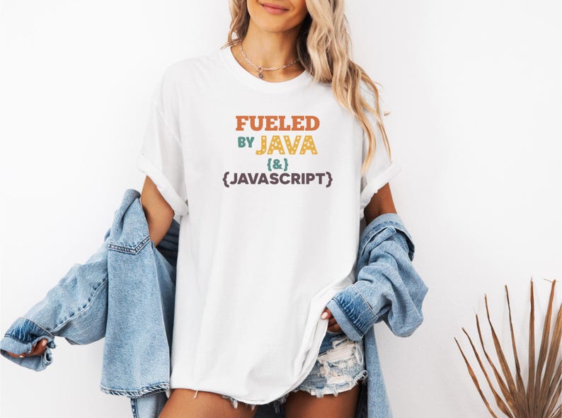 Op de afbeelding: Wit T-shirt met de tekst "FUELED BY JAVA (&) {JAVASCRIPT}" in oranje, groen, geel en paars. Het shirt wordt gedragen met een spijkerjasje en een spijkerbroek. Het algemene thema is tech en coderen.
