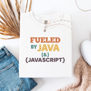 Op de afbeelding: Wit T-shirt met de tekst "FUELED BY JAVA & JAVASCRIPT" in oranje, groen, geel en paars. Het shirt is opgevouwen en ligt op een wit oppervlak naast een blauwe jeans, witte schoenen en een ketting.