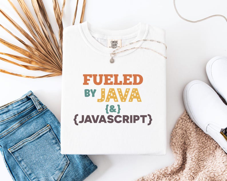 Op de afbeelding: Wit T-shirt met de tekst "FUELED BY JAVA & JAVASCRIPT" in oranje, groen, geel en paars. Het shirt is opgevouwen en ligt op een wit oppervlak naast een blauwe jeans, witte schoenen en een ketting.