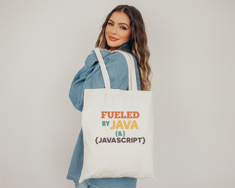 K&ouml;nnte beinhalten: Eine cremefarbene Tragetasche mit dem Text "FUELED BY JAVA {&} JAVASCRIPT" in Orange, Gr&uuml;n, Gelb und Lila. Die Tasche hat zwei cremefarbene Tr&auml;ger und wird &uuml;ber der Schulter einer Person getragen.