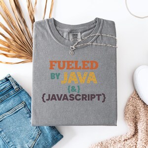 Op de afbeelding: Een grijs Comfort Colors T-shirt met de tekst "Fueled by Java & Javascript" in oranje, geel en paars. Een zilveren ketting met een hanger ligt op het shirt. Het shirt ligt naast een blauwe jeans, witte sneakers en een beige gebreid item.