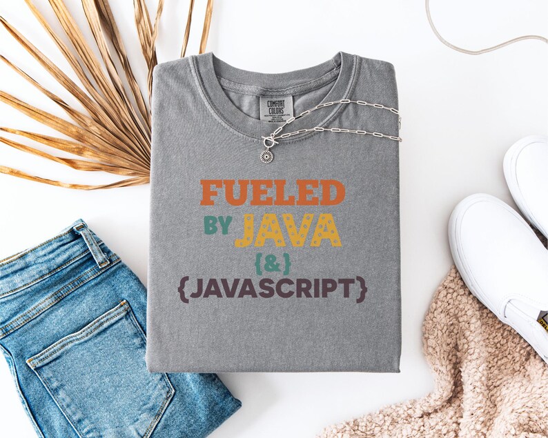 Op de afbeelding: Een grijs Comfort Colors T-shirt met de tekst "Fueled by Java & Javascript" in oranje, geel en paars. Een zilveren ketting met een hanger ligt op het shirt. Het shirt ligt naast een blauwe jeans, witte sneakers en een beige gebreid item.